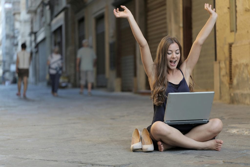 freelancer WordPress, A cheerful woman sitting outdoors, celebrating success with arms raised, while using a laptop.